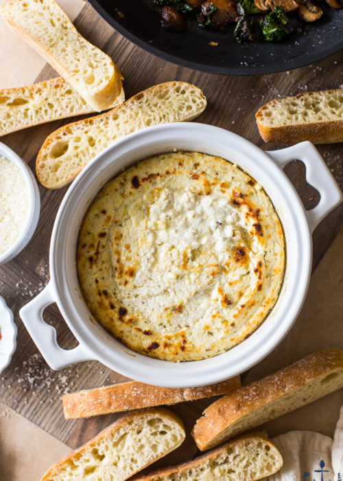 Overhead photo of Baked ricotta in a white crock on a wooden board surrounded by bread slices