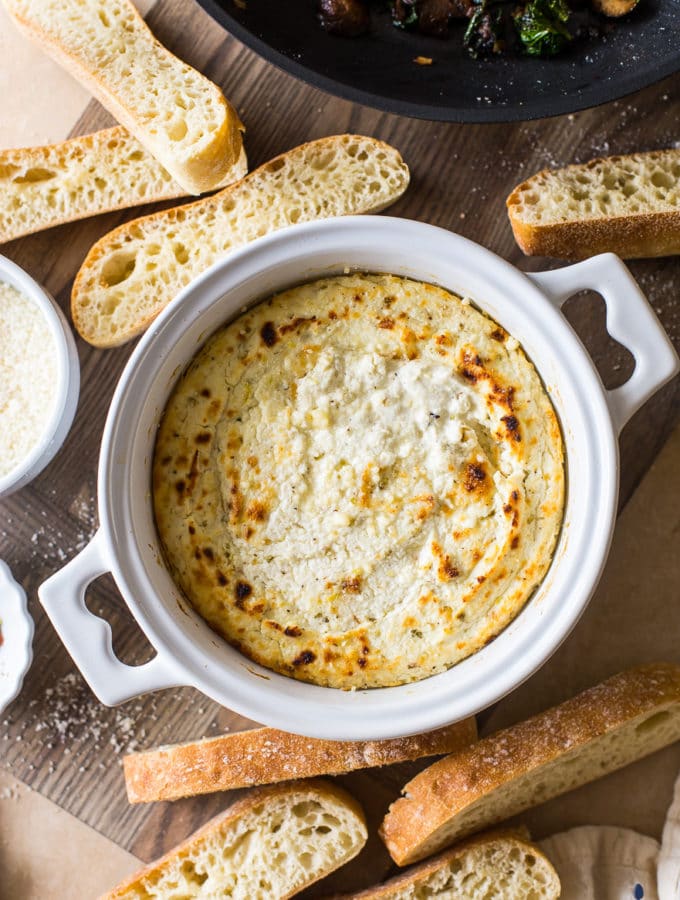 Overhead photo of Baked ricotta in a white crock on a wooden board surrounded by bread slices