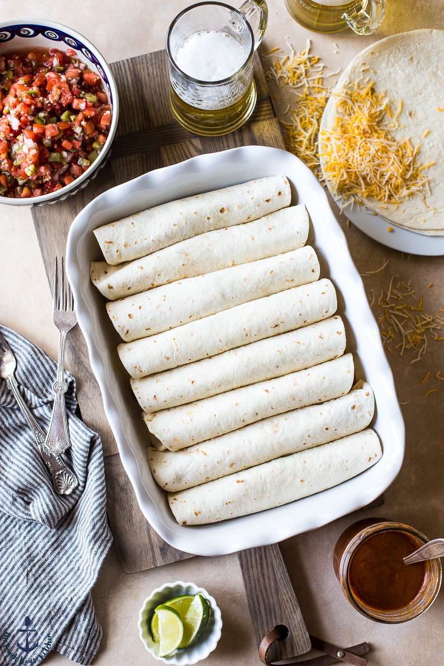 Prebaked enchiladas in a white dish with plenty of delicious sides around, including cheese, lime and salsa