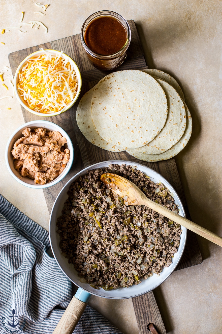 Ground beef mixtire in skillet, tortillas, shredded cheese and refried beans in bowls ready on a wooden board
