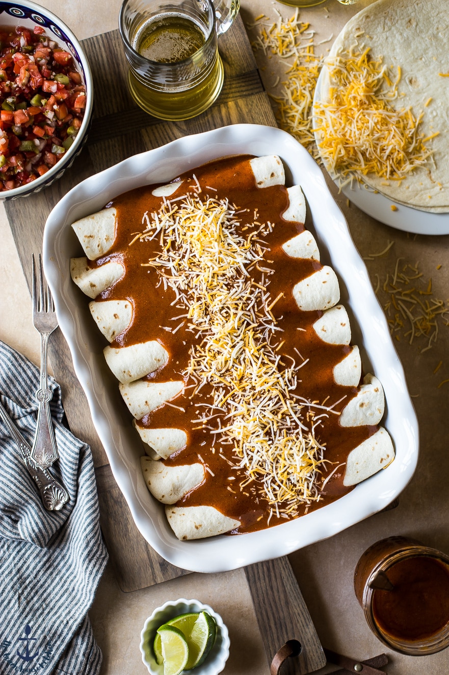 Overhead photo of prebaked enchiladas in a white dish topped with enchilada sauce and then - cheese