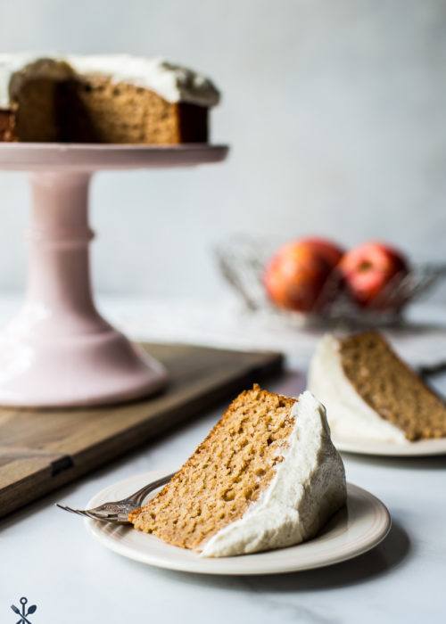 A slice of applesauce cake with vanilla bean frosting a a plate with a cake stand in background
