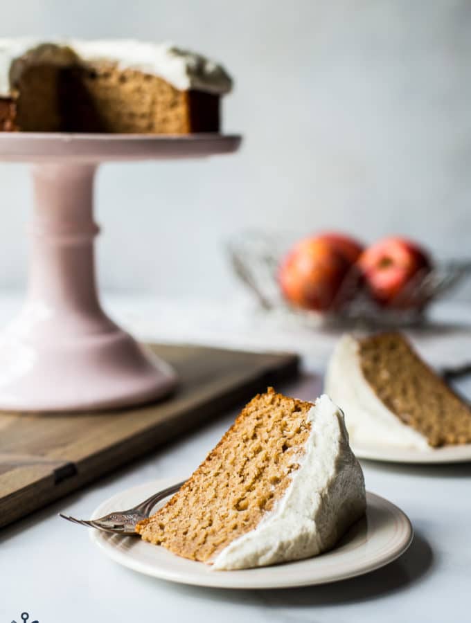 A slice of applesauce cake with vanilla bean frosting a a plate with a cake stand in background