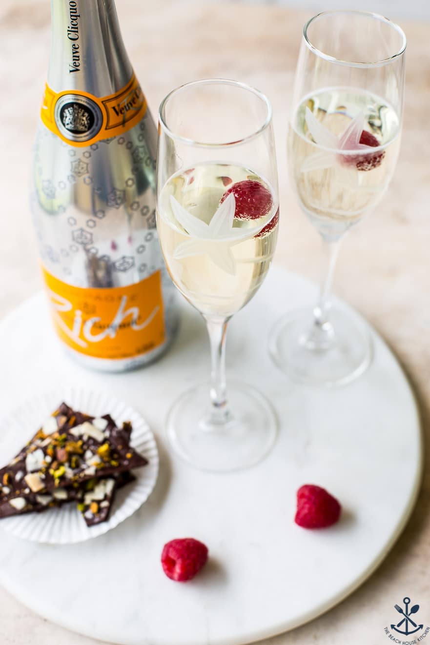 A round marble board with a bottle of champagne, two champagne glasses, raspberries and chocolate coconut pistachio bark