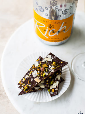 Chocolate coconut pistachio bark on a round marble board with a bottle of champagne
