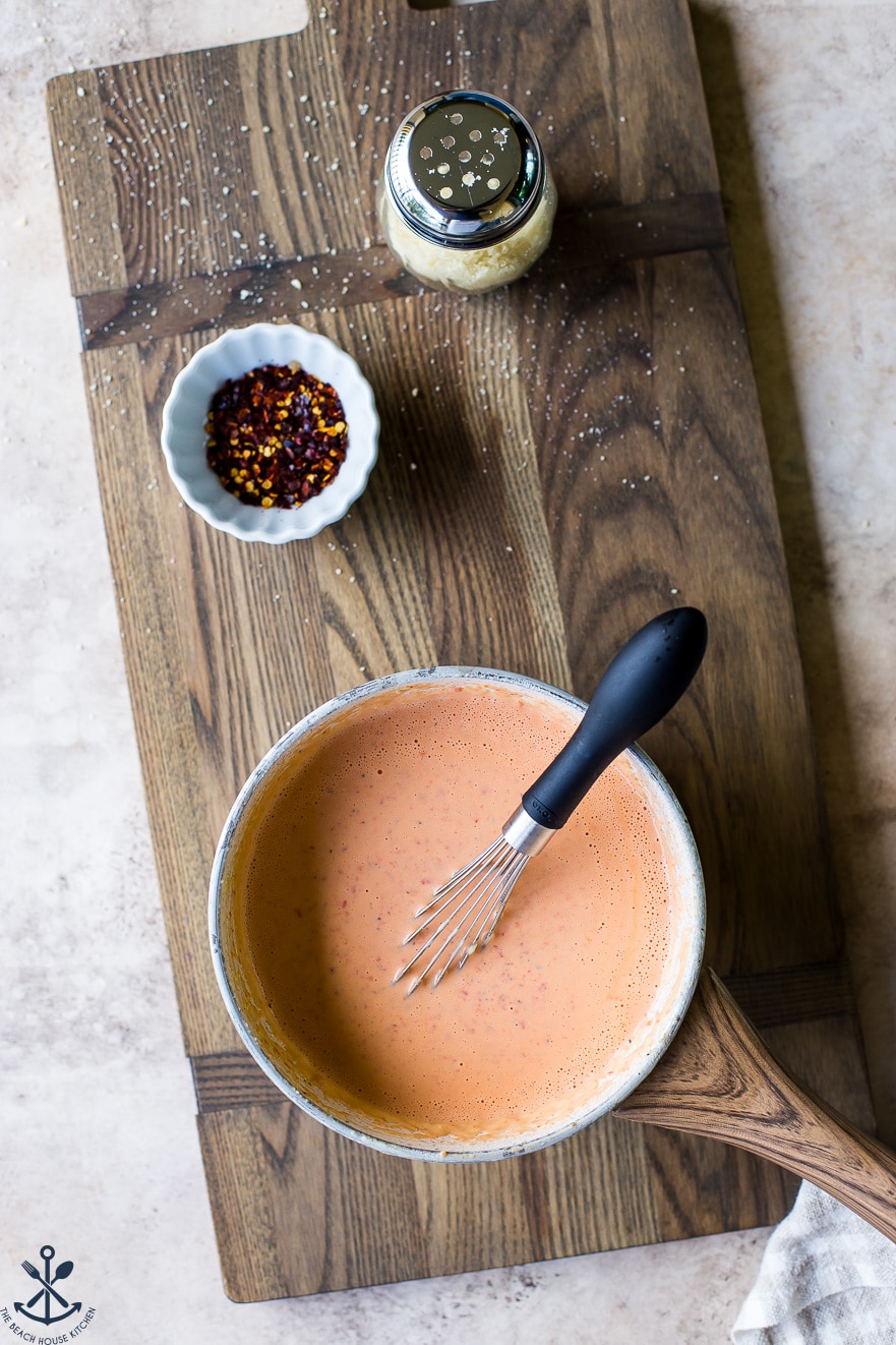 Overhead photo of roasted red pepper Alfredo sauce in a saucepan with a whisk on a wooden board