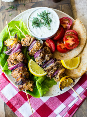 Overhead photo of Chicken Souvlaki Skewers with Tzatziki Sauce on a plate on top of a red and white checked napkin.
