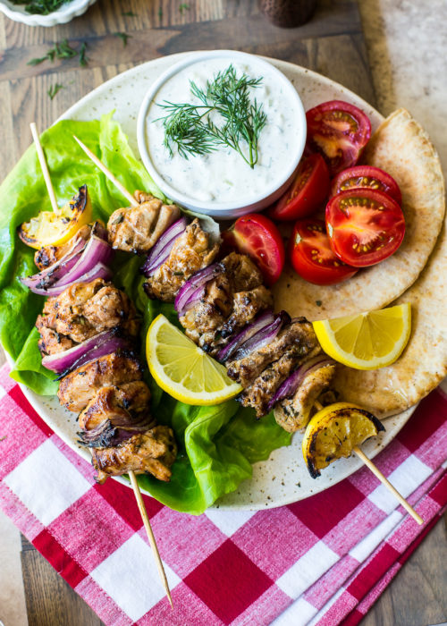Overhead photo of Chicken Souvlaki Skewers with Tzatziki Sauce on a plate on top of a red and white checked napkin.