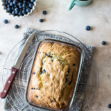 Overhead photo of blueberry lemon loaf on a round wire rack with a bowl of blueberries and a cup of coffee