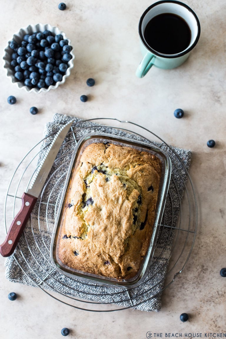 Overhead photo of blueberry lemon loaf on a round wire rack with a bowl of blueberries and a cup of coffee