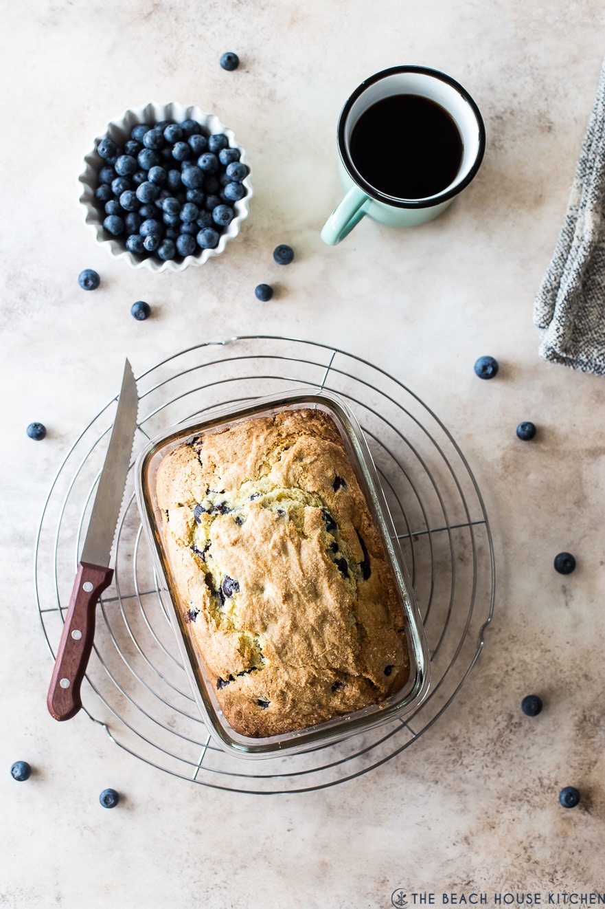 Blueberry lemon loaf on a round wire rack with a bowl of blueberries and a cup of coffee next to the rack