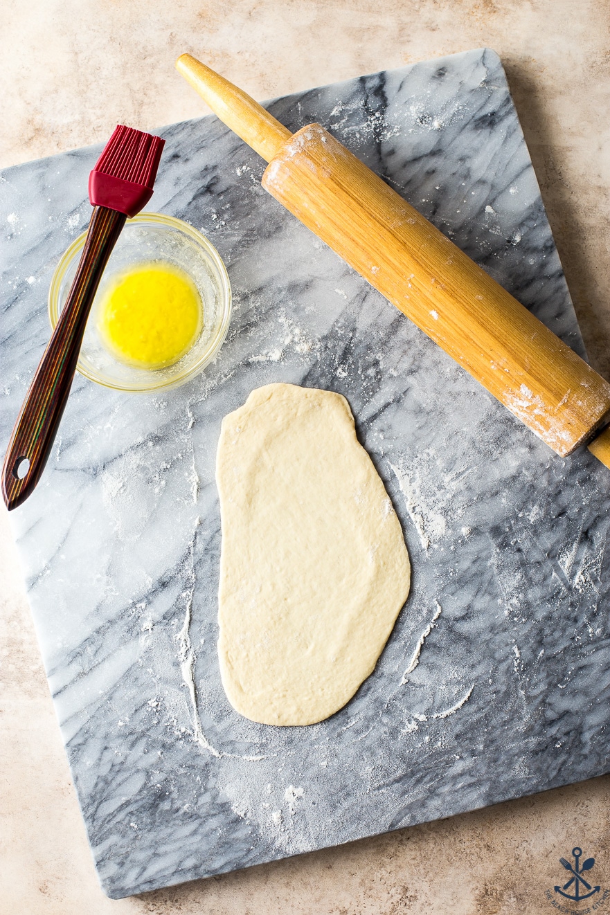Overhead photo of naan dough rolled into an oval on a marble board with a rolling pin, brush and bowl of melted butter