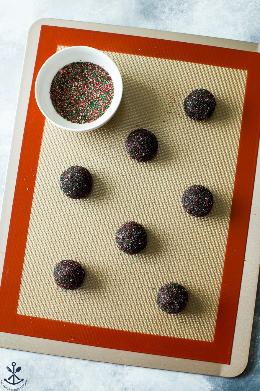 Prebaked cookie balls on a baking sheet lined with a silicone baking mat with a bowl of colored sugar