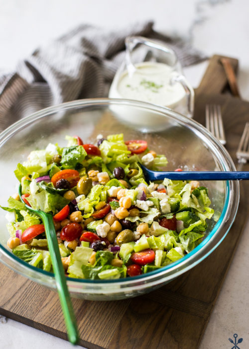 Greek Chickpea Salad with Green Goddess Dressing in a bowl on a wooden board with dressing in a glass pitcher in the background