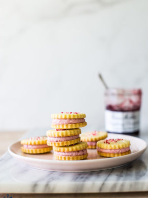 Key Lime Sandwich Cookies with Raspberry Buttercream on a pink plate on a marble board with a jar of preserves in the background