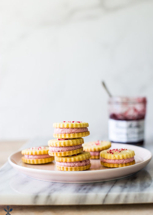 Key Lime Sandwich Cookies with Raspberry Buttercream on a pink plate on a marble board with a jar of preserves in the background