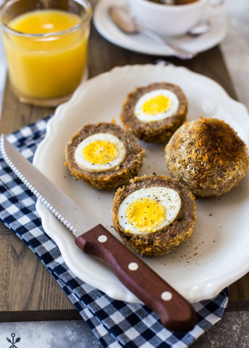 A plate with scotch eggs sliced in half with a sharp knife on a blue and white checked napkin