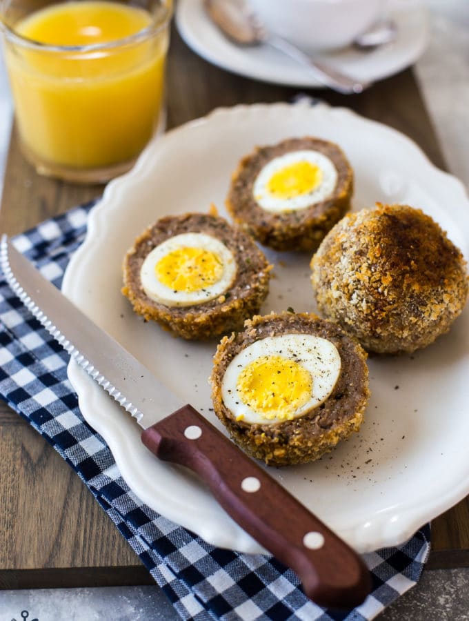 A plate with scotch eggs sliced in half with a sharp knife on a blue and white checked napkin
