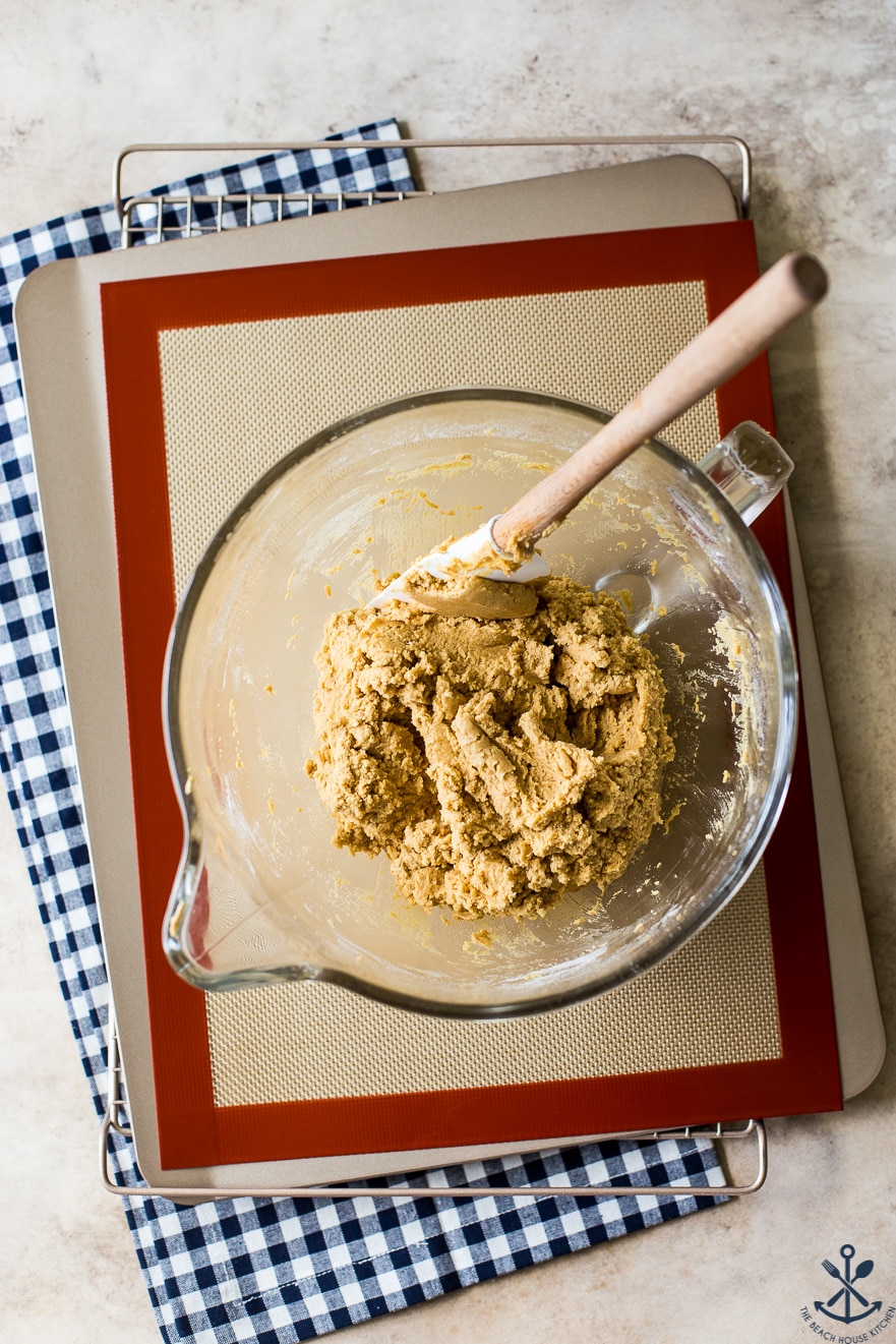 Overhead photo of bowl filled with peanut butter cookie dough on a silpat lined baking sheet on a blue and white checked napkin