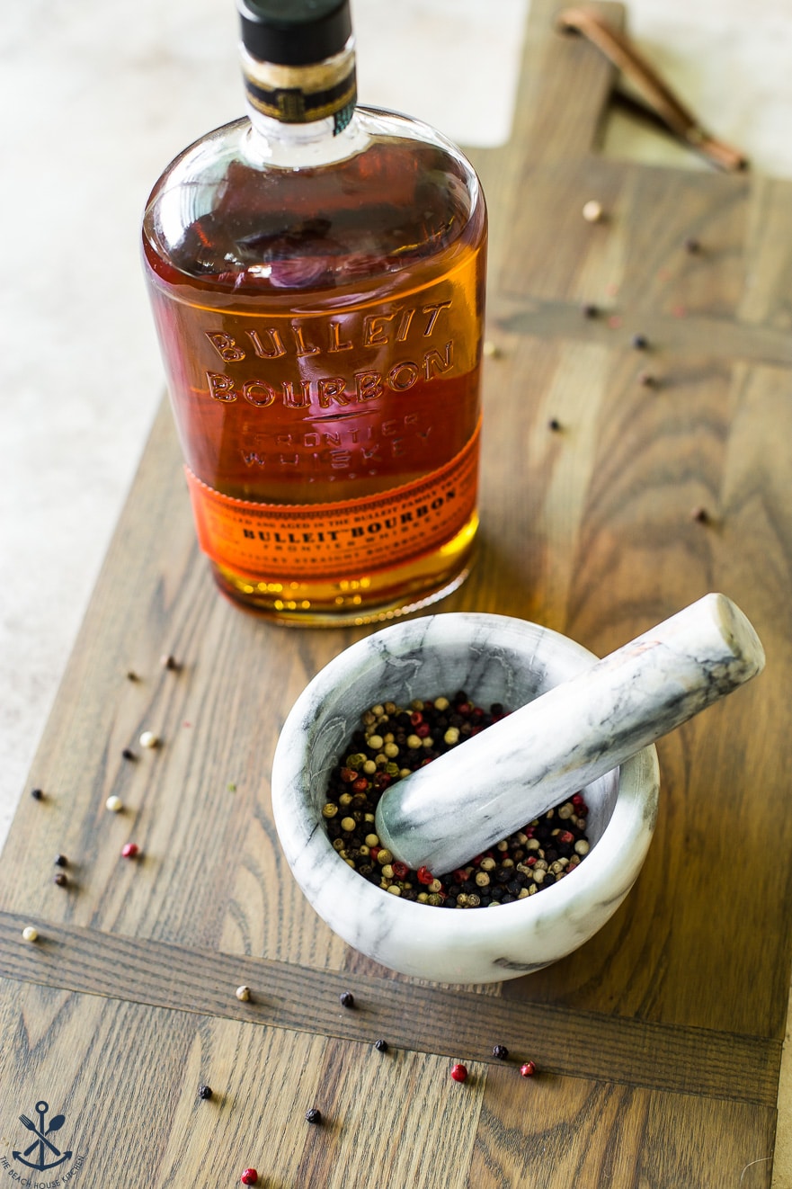 Bowl filled with peppercorns and a bottle of bourbon on a wooden board next to it