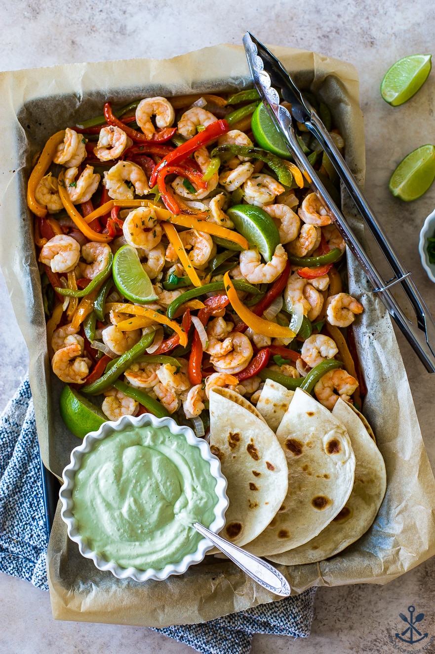 Overhead photo of a large sheet pan with shrimp, peppers, onions and a bowl of avocado crema