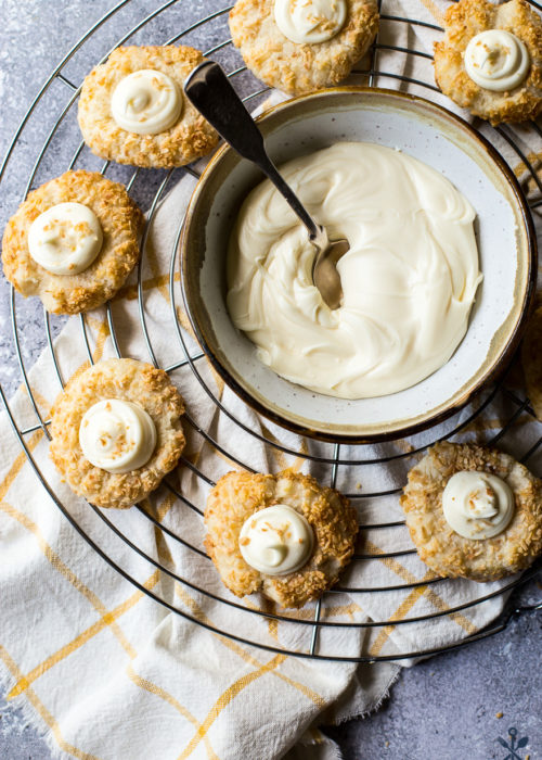 Overhead photo of round baking rack with coconut thumbprint cookies and a bowl of cream cheese frosting with a spoon