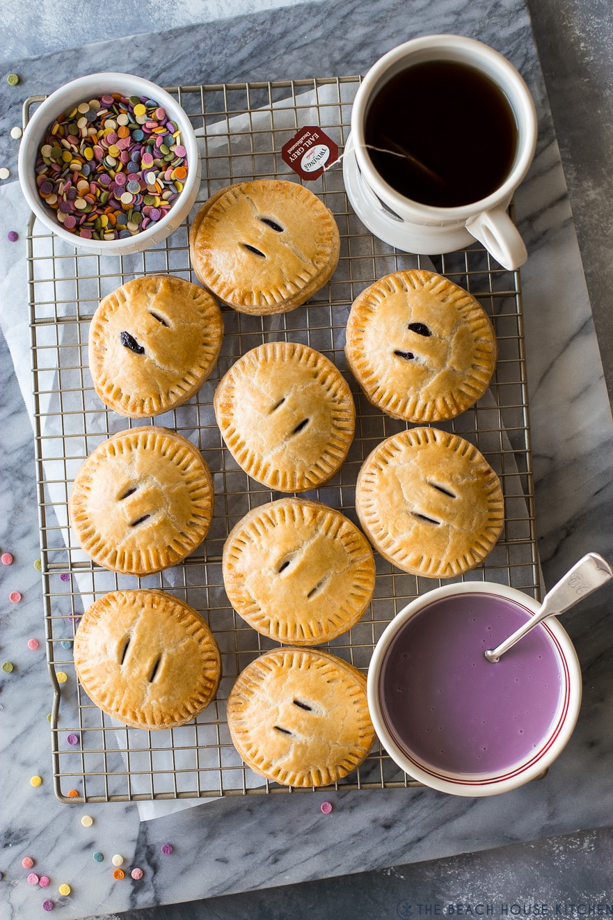 Overhead photo of round hand pies on a wire rack with a bowl of sprinkles, purple glaze and a mug of tea