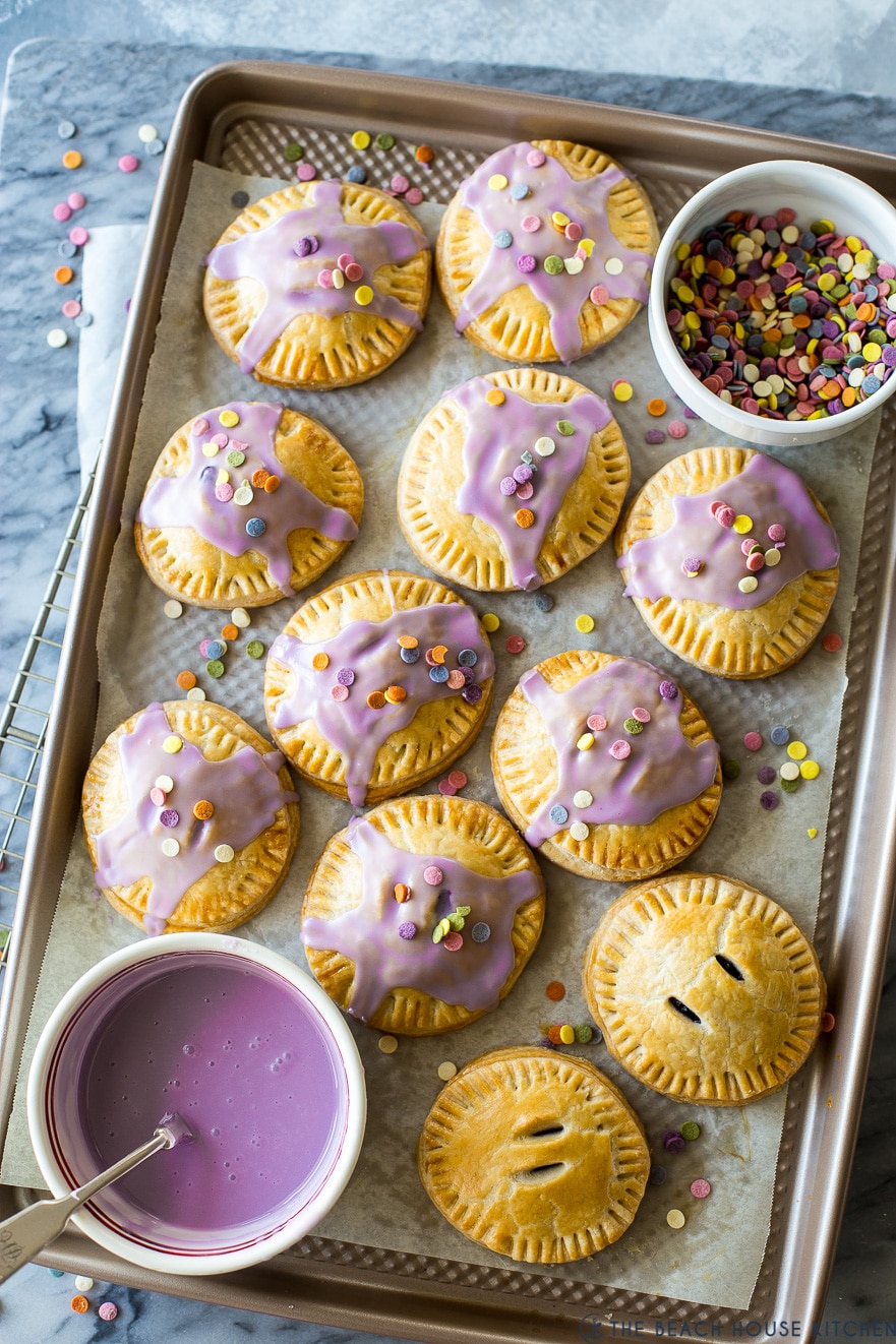 Overhead photo of a baking sheet filled with purple glazed hand pies and a bowl of purple glaze and sprinkles