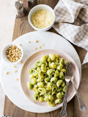 Overhead photo of a pink plate of Broccoli Pesto Pasta on a round marble board with a small bowl of pine nuts