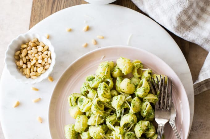 Overhead photo of a pink plate of Broccoli Pesto Pasta on a round marble board with a small bowl of pine nuts