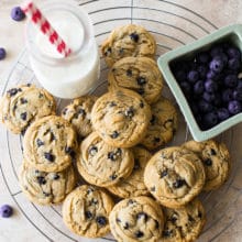 Overhead photo of Chewy Brown Sugar Blueberry Cookies on a round wire rack with a bottle of milk and a basket of blueberries