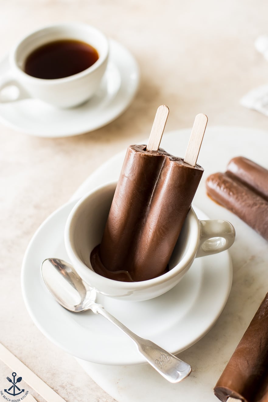 Up close photo of a creamy mocha popsicle in a coffee cup with a silver spoon and a cup of coffee in the background