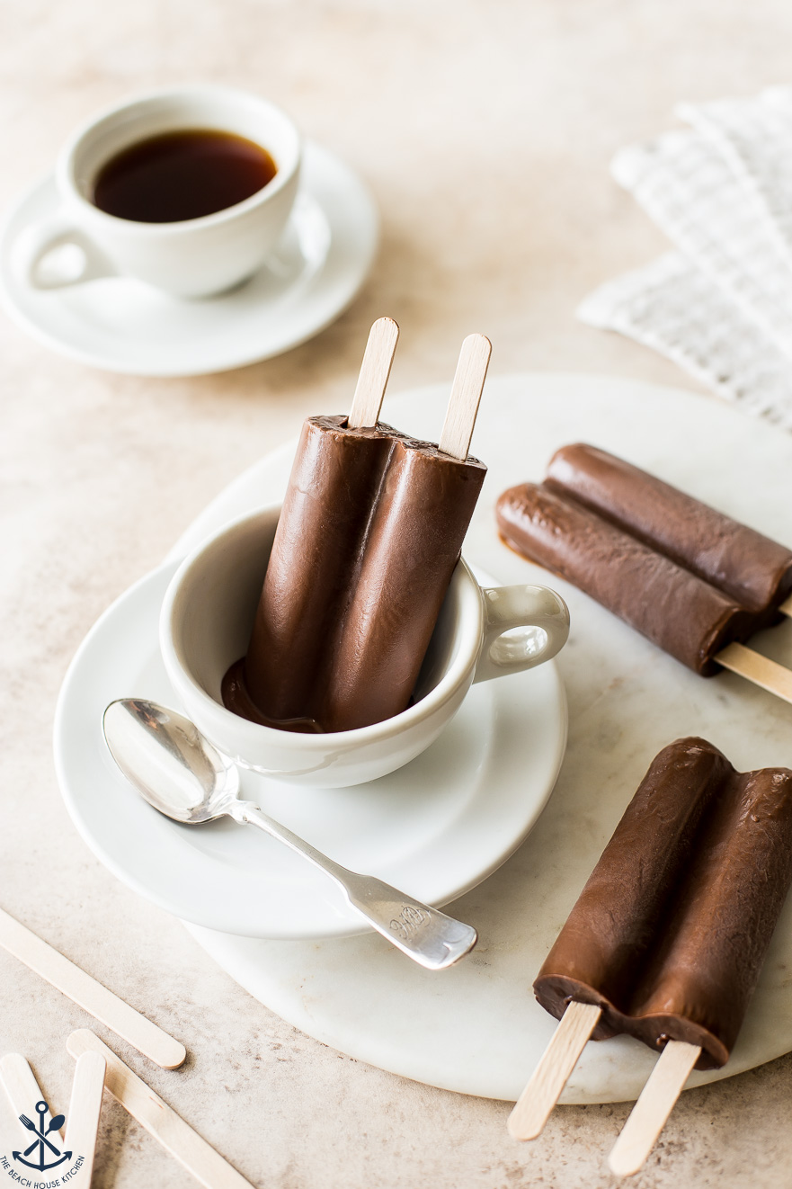 A chocolate popsicle in a coffee cup with two popsicles off to the side and a coffee cup in the background