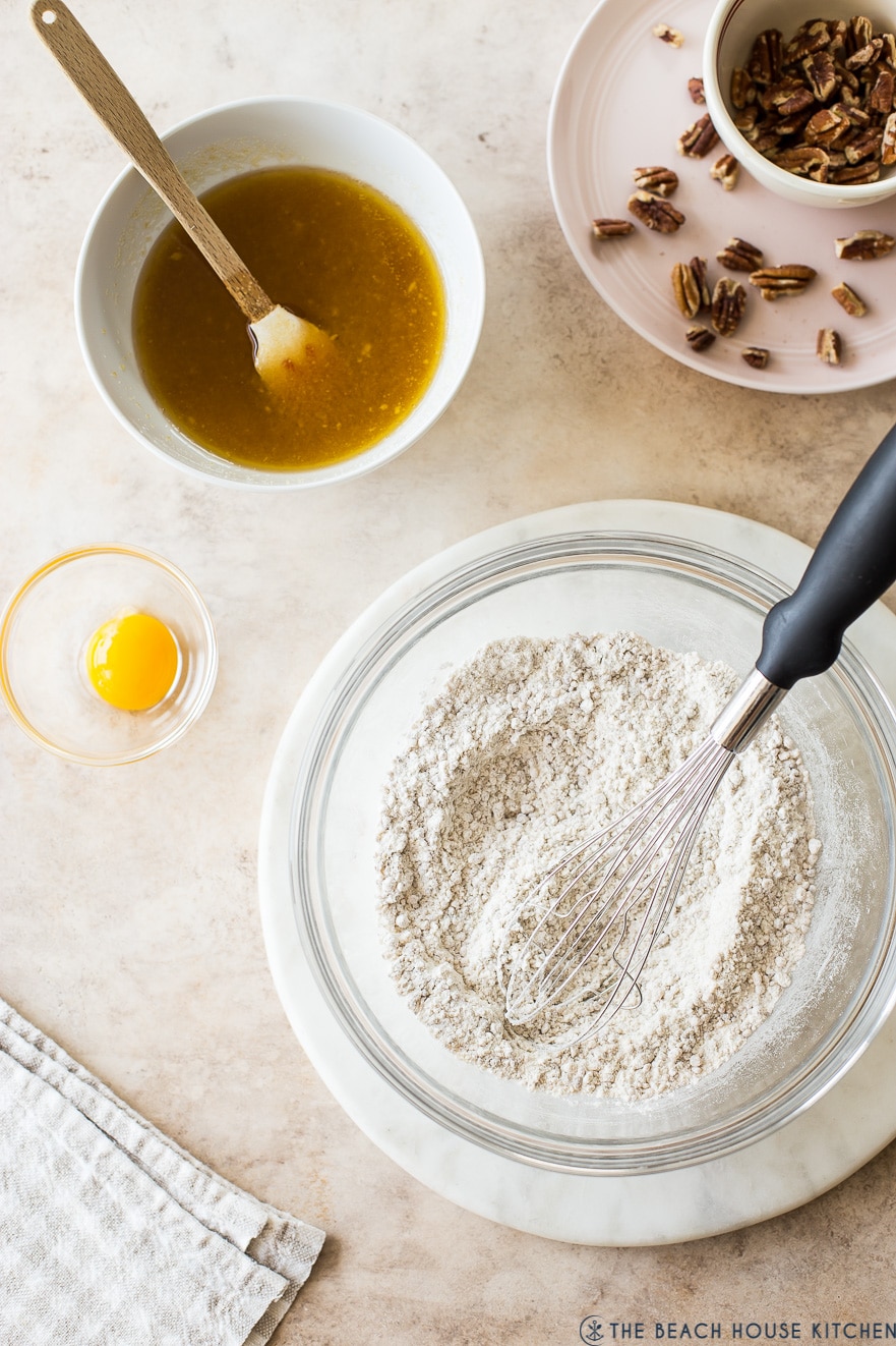 Overhead photo of flour in a bowl with a whisk with a bowl of melted butter and brown sugar in a white bowl and an egg yolk in a small bowl