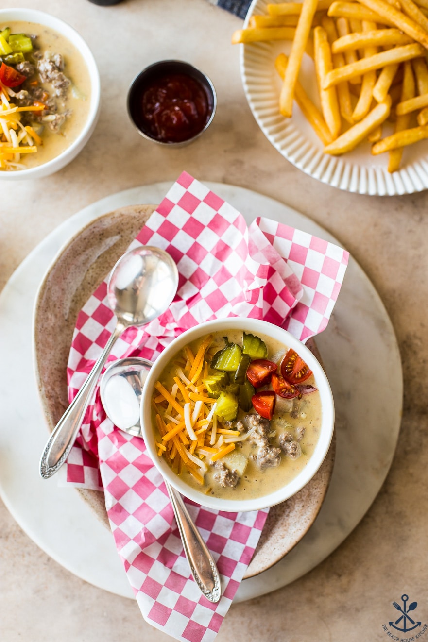 Overhead photo of a bowl of cheeseburger soup on a plate with a red and white check paper and two spoons