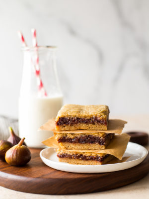A stack of three fig bars on a white plate with a bottle of milk in the background