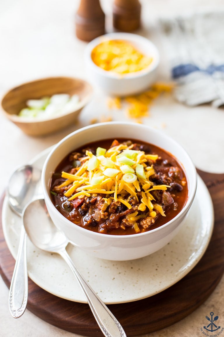 A white bowl filled with turkey chili on a white plate with two spoons