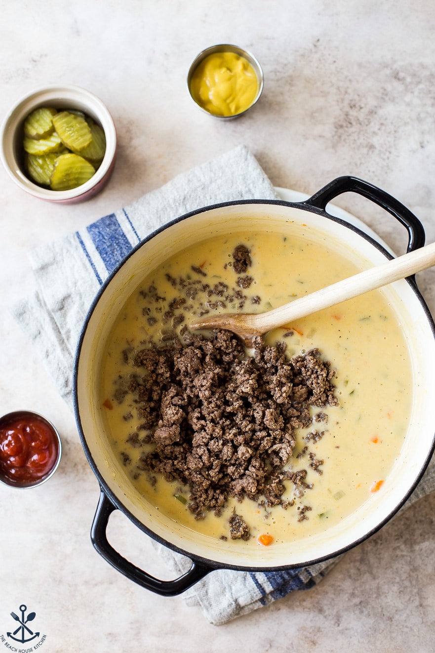 Overhead photo of a pot of soup topped with cooked ground beef with a bowl of pickles off to the side 