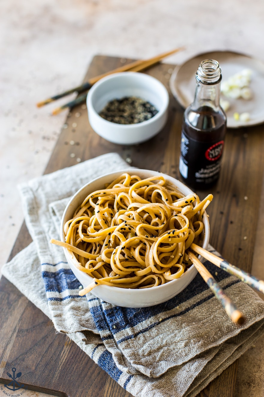 A bowl of sesame noodles with chopsticks in a white bowl on a wooden board 