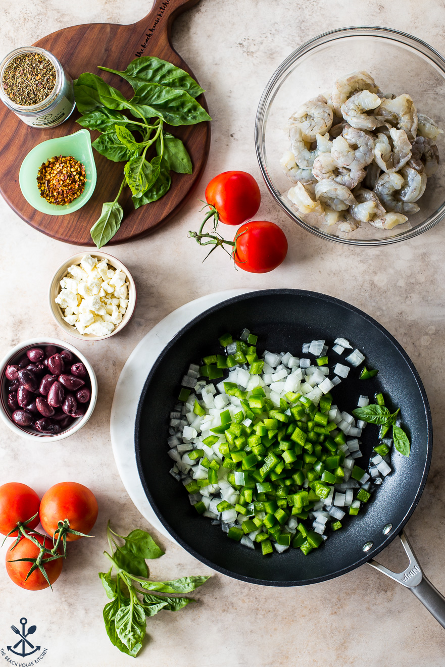 Overhead photo of a skillet filled with chopped green peppers and onions surrounded by a bowl of raw shrimp, a bowl of Kalamata olives and a bowl of feta crumbles