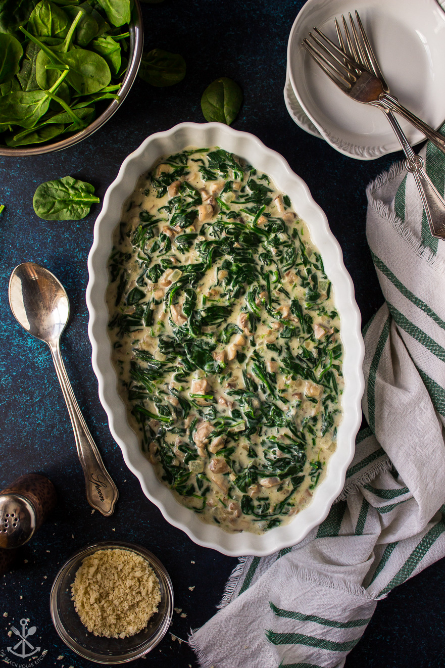 Overhead photo of an oval white dish filled with creamed spinach on a blue backgrounds with a spoon off to the side