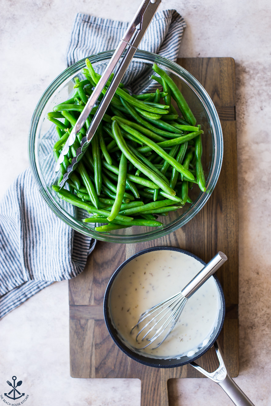 Overhead photo of a bowl of green beans and a small pot of white sauce with a whisk
