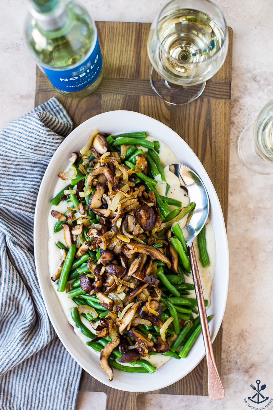 Overhead photo of an oval plate of green beans and mushrooms on a wooden board with a bottle and glass of white wine
