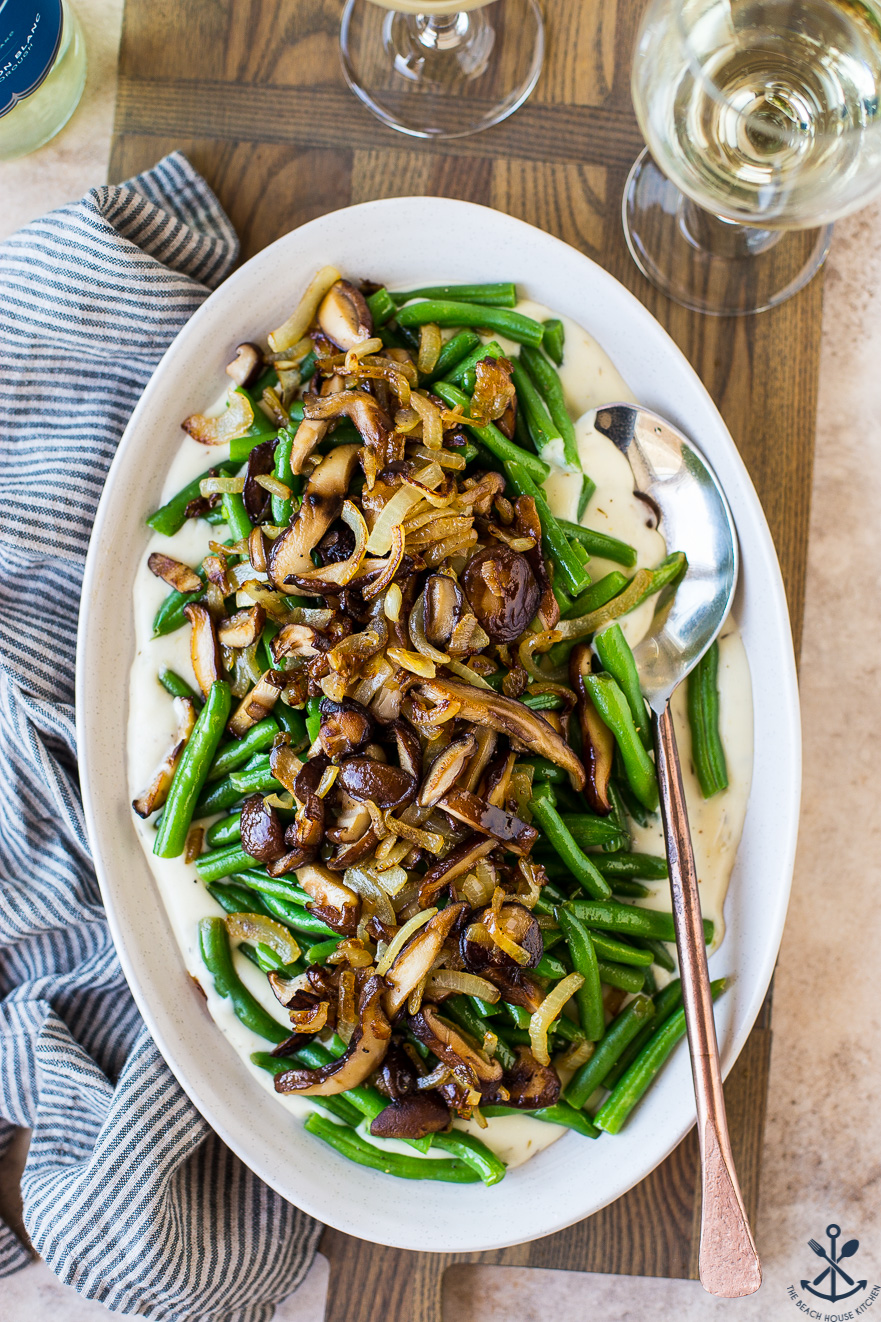 Up close overhead photo of an oval plate of green beans topped with mushrooms with a spoon and a glass of white wine