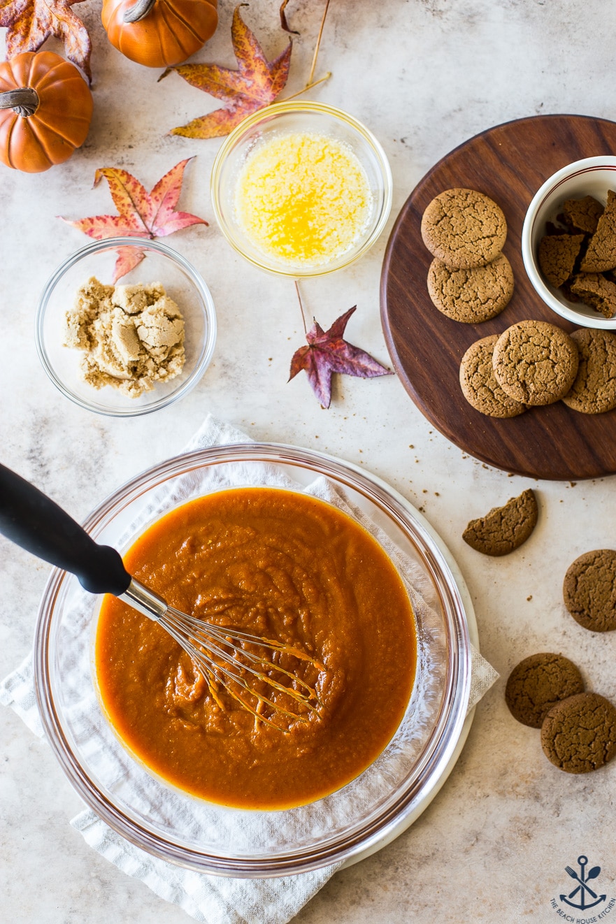 Overhead photo of bowl filled with pumpkin mixture and a whisk and a dark wood board to cookies