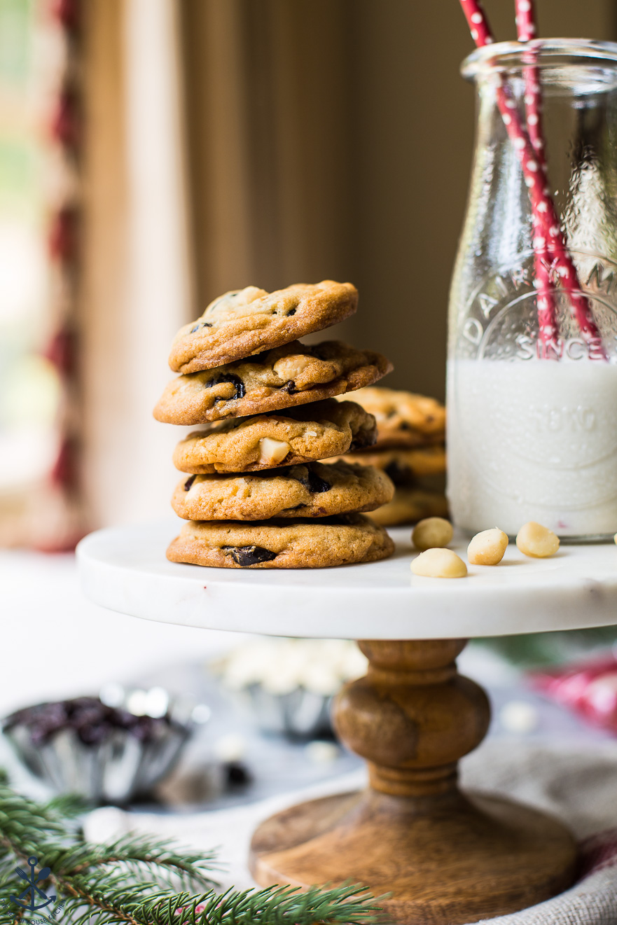 Up close photo of a cake stand topped with a stack of white chocolate cherry macadamia nut cookies and a bottle of milk with two red straws