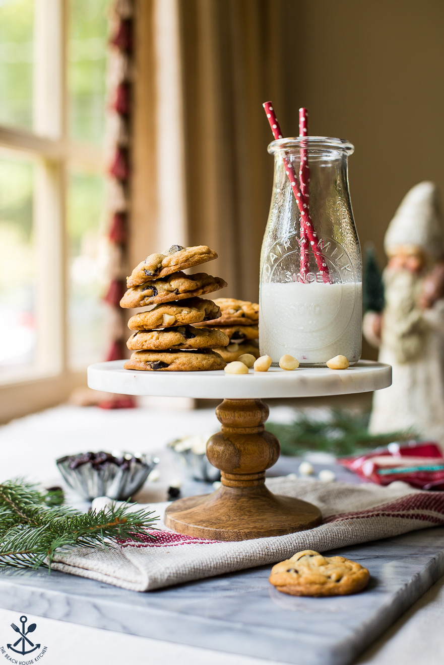 A cake stand topped with a stack of white chocolate cherry macadamia nut cookies and a bottle of milk with two red straws