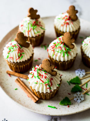 A plate of cream cheese frosted cupcakes with red and green sprinkles topped with a gingerbread man cookie