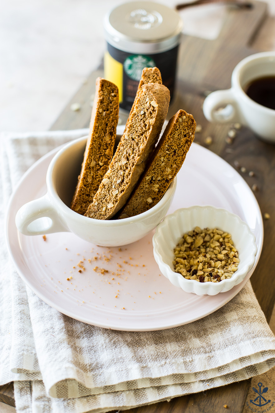 Slices of pre-chocolate dipped hazelnut biscotti in a coffee cup with a small bowl of hazelnuts all on a pink plate