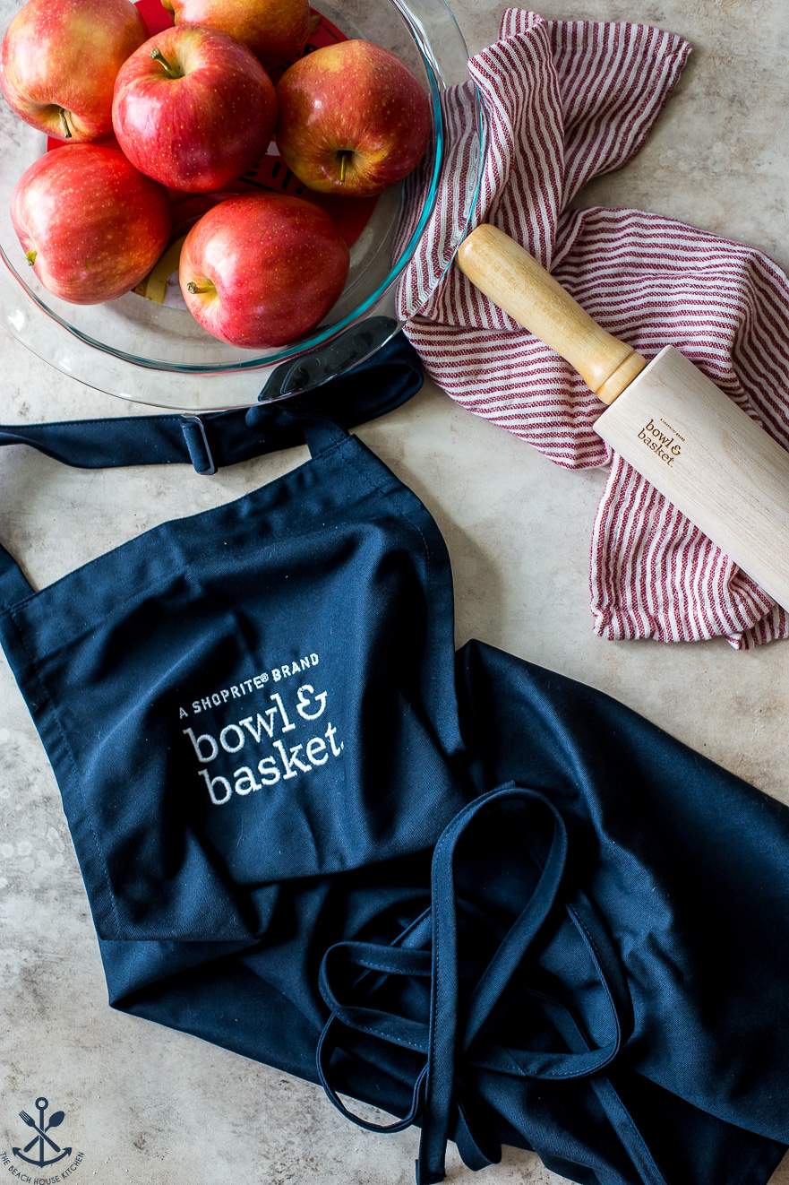 Overhead photo of a navy blue Bowl & Basket apron with a pie plate filled with apples and a rolling pin off to the right side